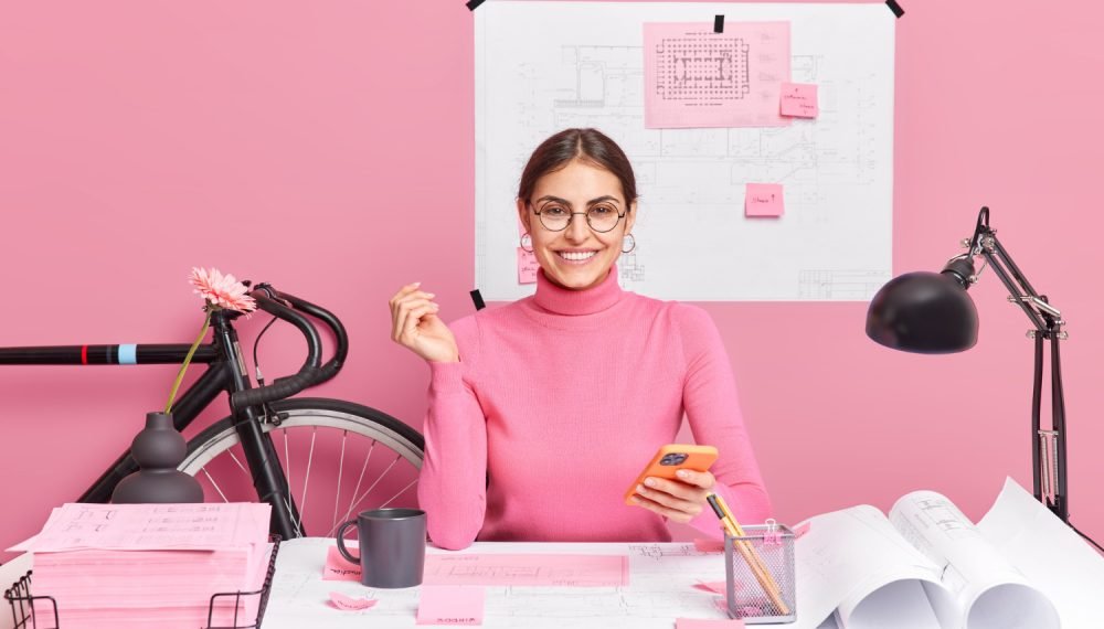 cheerful-female-graphic-designer-holds-modern-smartphone-creats-blueprint-drawing-sketch-poses-desktop-wears-spectacles-turtleneck-sits-coworking-space-successful-architect-woks-project-1000x570